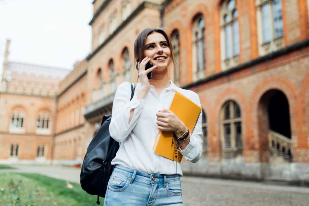 Smiling college student standing outside a university building, talking on the phone and holding notebooks, representing student success, financial aid support, FAFSA guidance, and continuous improvement for colleges in Texas focused on compassionate and proactive student support.